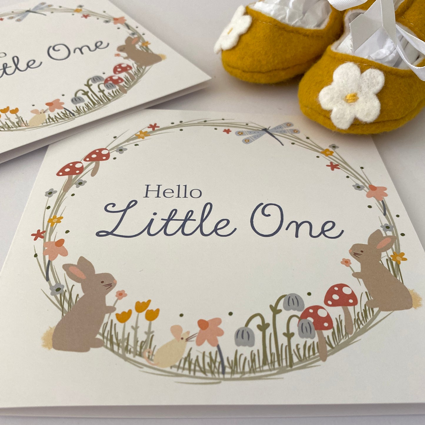 Hello Little One new baby greeting card hand drawn by Kathrin Legg. A beautiful, soft pastel woodland wreath illustratoin featuring small baby rabbits, mushrooms and a dragon fly with the words 'Hello Little One' featured in the centre of the wreath.