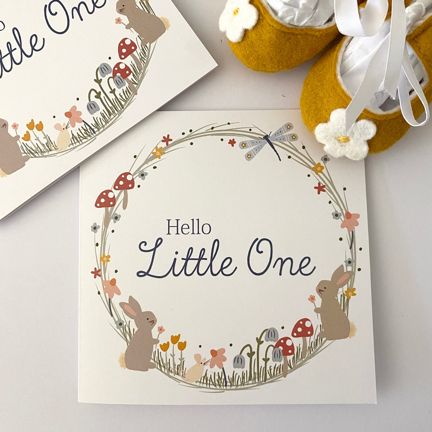 Hello Little One new baby greeting card hand drawn by Kathrin Legg. A beautiful, soft pastel woodland wreath illustratoin featuring small baby rabbits, mushrooms and a dragon fly with the words 'Hello Little One' featured in the centre of the wreath. Photographed by Kathrin Legg with a pari of baby felt shoes.